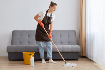 Fototapeta premium Full length portrait of woman in good mood wearing brown apron, jeans and white t shirt cleaning her house, washing floor near sofa, likes doing household chores.
