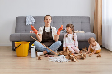 Indoor shot of woman wearing brown apron, jeans and white t shirt cleaning her house with children...