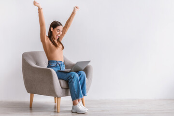Excited young Caucasian lady sitting in armchair with laptop, lifting hands up, celebrating success...