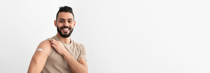 Cheerful Arab man showing arm with band aid after receiving covid vaccine injection, smiling,...