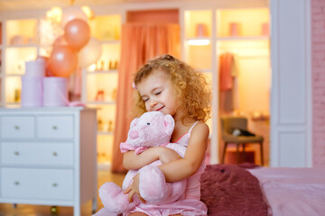 Fototapeta premium Portrait of a small curly blonde with a stuffed toy in the bedroom. A girl hugs a pink bear.