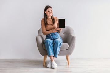 Happy woman showing tablet with empty screen, sitting in armchair against white wall, mockup for...