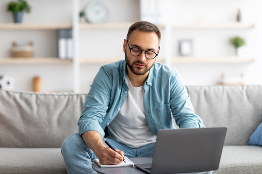 Serious Young Man Using Laptop, Working Online, Taking Notes During Business Meeting From Home Office