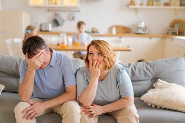 Tense parents sitting on sofa tired crazy loud hyperactive little kids, siblings at home. An...