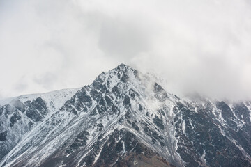 Awesome landscape with high snowy mountain peaked top with sharp rocks in low clouds. Dramatic view to snow mountain pointed peak in low cloudy sky. Monochrome scenery with white snow on black rocks.