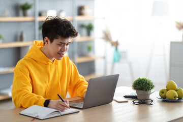 Smiling asian man working on laptop and writing