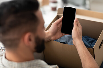 Unrecognizable young Arab guy holding smartphone with mockup for app or website on screen while unboxing parcel at home