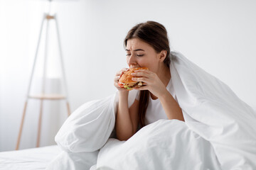 Hungry after diet, starving stressed caucasian millennial woman eating burger in bed