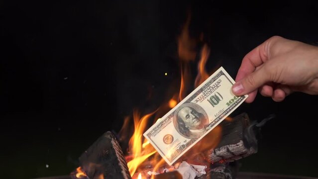 hand puts dollars burn in fire camp. US dollars banknotes on fire. Black background, isolate. concept of inflation, decrease in the purchase of foreign currency and slow motion paper die