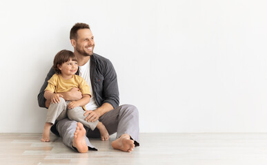 Happy caucasian father carrying little son, sitting together on floor and looking aside at empty...