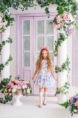 girl who looks like a doll is standing on the veranda of a house decorated with flowers. A child with long blonde curls and a big pink bow on her head