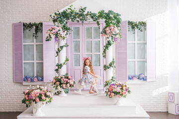 girl is sitting on a wooden rocking horse on the veranda of a house decorated with flowers. A child with long blonde curls and a big pink bow on her head