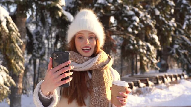Portrait of happy young woman talking on video call via mobile phone and holding cup with hot coffee in sunny winter cold day. Smiling cute female in warm clothes standing on snowy street, slow motion