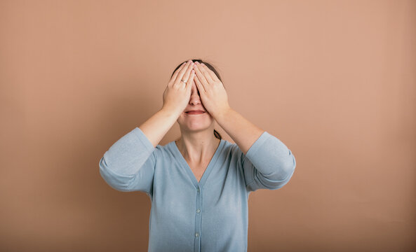 a brunette woman with glasses in a blue jumper stands and covered her eyes with her hands on a beige background with a place for text