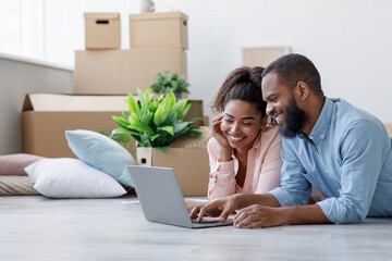 Smiling millennial black couple rest, lie on floor with cardboard boxes with things and plants in...