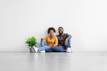 Happy millennial black couple sit on floor with plant in pot on gray wall background in empty room, copy space