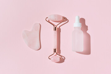 Facial kit for home skin care and spa. Face roller, gua sha massager and bottle of cosmetic serum on pink pastel sunlit background with shadows. Natural treatment concept. Top view. Flat lay
