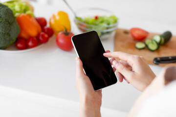 Healthy food and diet planning concept. Young woman using smartphone with blank screen, preparing salad in kitchen