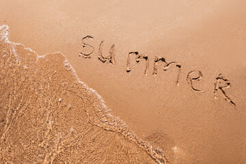 Summer text is written on the beach yellow sand with a wave near the sea, top view. Summer vacation and rest concept