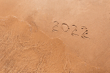 Happy New Year 2022, lettering on the beach with wave and clear sea. Numbers 2022 year on the sea shore, message handwritten in the golden sand on beautiful beach background. New Years concept.