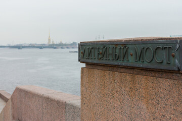 Obraz premium Close-up view of parapet fragment with name of Liteyny Bridge at cold cloudy day, defosused Neva panorama at background. Bronze and granite. Copy space for text. St.Peterburg, Russia
