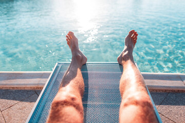 Male tanned legs lie on a beach lounger on a background of a swimming pool with blue water. Summer...