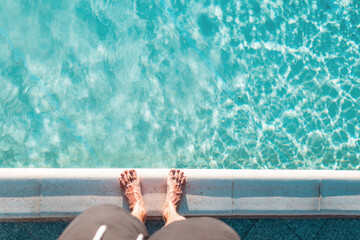 Fototapeta na wymiar Male feet stand near the swimming pool with blue beautiful water on a sunny day, top view. Summer vacation concept