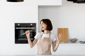 Young happy housewife dancing and singing a song in modern kitchen interior, pretending whisk to be...