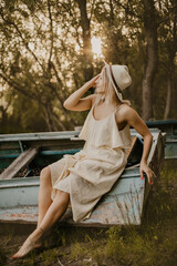 Fototapeta na wymiar blonde girl in a light dress and a hat near an old boat on the beach