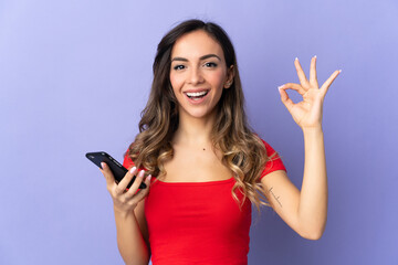 Young caucasian woman isolated on purple background using mobile phone and doing OK sign