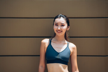 Portrait of Smiling Asian Woman in Sportswear Standing In Front of Wooden Wall. 
Confident female athlete looking at camera while taking a break of outdoor workout training.