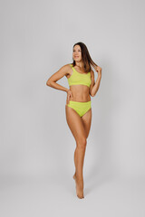 Brunette shows off slender figure. Young beautiful Caucasian woman in yellow swimsuit or bikini poses on light isolated background, stands in elegant pose and holds her hair with her hands. 