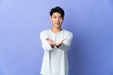 Young Chinese man isolated on purple background holding copyspace imaginary on the palm to insert...