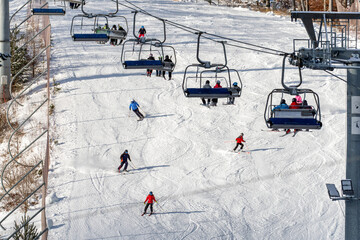People sitting on chairlift and skiers skiing downhill on slope at mountain resort.
