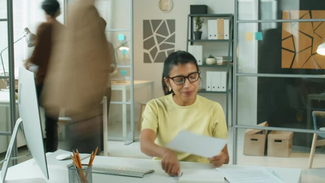 Time lapse of young Hispanic businesswoman working at desk in office. She using computer, taking notes, reading documents and cooperating with multiethnic colleagues