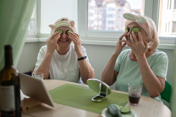 Two senior women apply facial cosmetic mask, have fun at home, make selfie and chat with friends in...