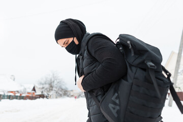 Fototapeta na wymiar Handsome fashionable man with a black textile mask in fashionable black clothes with a vest, a hat and backpack walks outdoors with snow. The guy travels and Journey during a pandemic and coronavirus