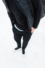 Men's winter black clothing style. Fashionable man in black vest, hoodies, pants, sneakers with a backpack stands in the snow