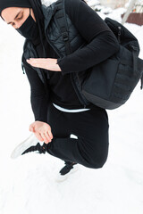 Fashionable man in stylish black winter clothes with a backpack posing in the snow. Guy in ninja style outerwear