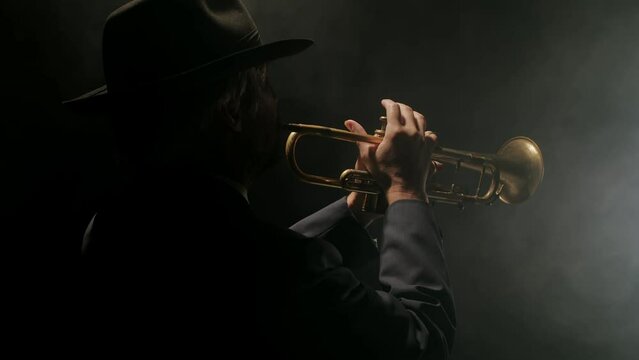 Jazzman plays trumpet in dark, smoky room in spotlight, back view. Man at suit and hat plays blues on dark background