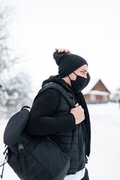 Handsome hipster man model with black mask in fashionable black outerwear with a black sports backpack walks and travels in countryside on a winter snowy day.