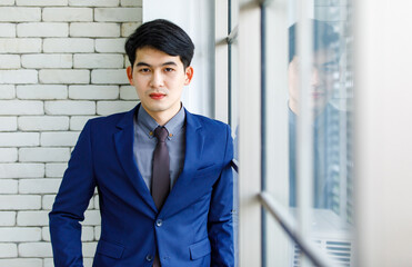 Portrait studio shot Asian handsome professional successful male businessman entrepreneur in formal blue suit necktie standing smiling look at camera on white brick wall background near office window
