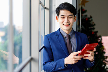 Fototapeta na wymiar Millennial Asian handsome smart confident professional success male manager businessman entrepreneur in formal blue suit smiling holding smartphone standing near skyscraper office window