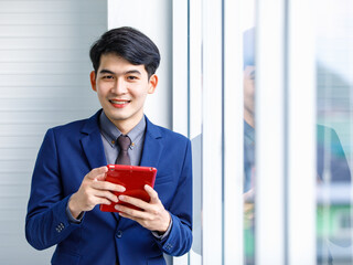Millennial Asian handsome smart confident professional success male manager businessman entrepreneur in formal blue suit smiling holding smartphone standing near skyscraper office window