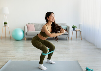 Fit young Indian woman doing squats with elastic band during home workout, copy space. Domestic...