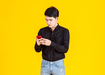 Studio shot of millennial Asian excited male businessman model in casual black long sleeve shirt and jeans standing holding red smartphone in hands playing virtual video game on yellow background