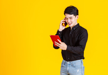 Studio shot of millennial Asian worried stressed male businessman model in casual black shirt and jeans standing talking on phone call with smartphone holding red tablet in hands on yellow background