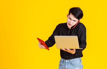 Portrait studio shot of Asian worried stressed male businessman in casual black shirt and jeans standing talking with smartphone holding red tablet and laptop computer in hands on yellow background