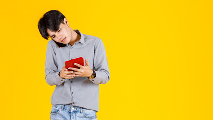 Studio shot of millennial Asian thoughtful doubtful curious male fashion model in stylish fashionable casual outfit standing call smartphone and holding red tablet on yellow background.