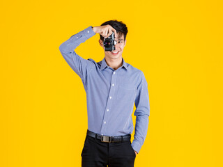 Fototapeta premium Portrait closeup studio shot of millennial Asian professional male fashion photographer journalist in casual outfit standing smiling holding aiming digital DSLR mirrorless camera on yellow background